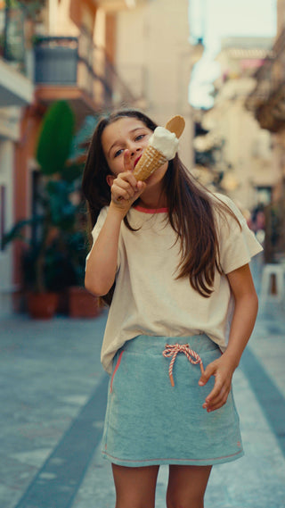 A young girl with long dark hair is shown from a low-angle perspective, wearing a white t-shirt and a light blue denim skirt while eating an ice cream cone outdoors.