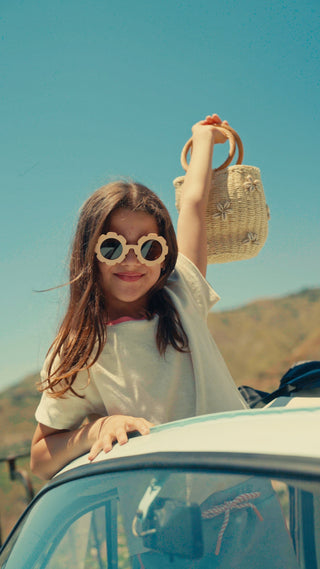 A girl leaning out of a car window against a bright blue sky, wearing round wooden-framed sunglasses, a white t-shirt, and holding a woven straw bag.