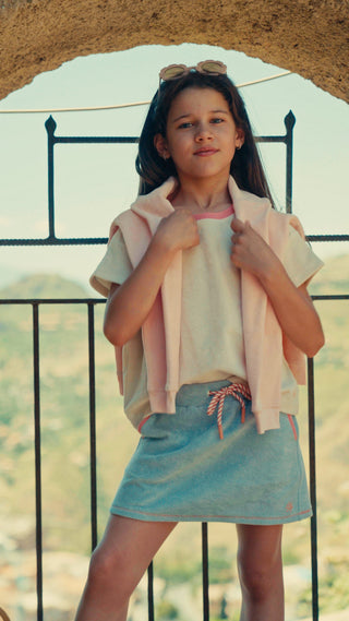 A young girl wearing a light blue denim skirt and a white t-shirt with a pink collar, holding a light pink cardigan. She is standing outdoors in front of a black metal railing with a sandy archway on the right.